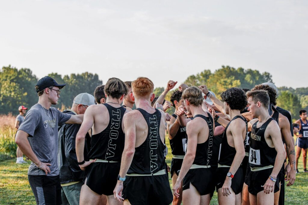 A group of male cross-country runners in black uniforms huddling together outdoors.