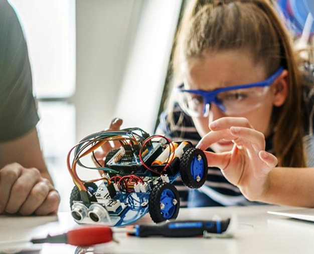 A person wearing safety goggles is focused on assembling a small wheeled robot, with various wires and electronic components visible.