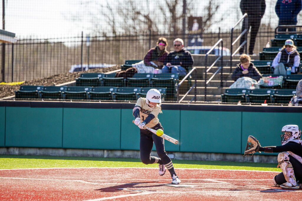 A softball player hitting a ball during a game, with spectators watching from the stands.