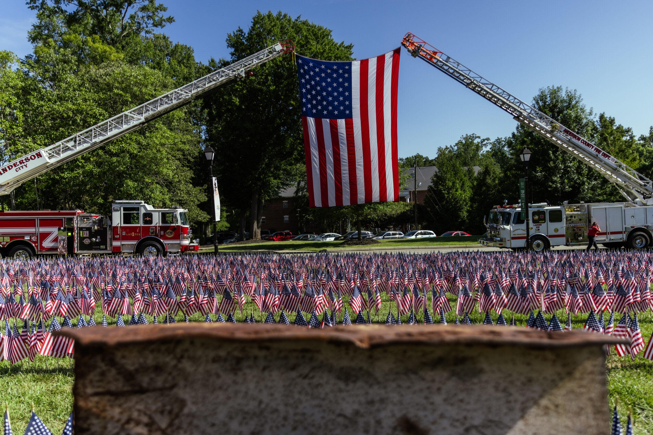 An American flag is displayed between two fire truck ladders above a field of American flags in the mid-ground and a remnant of the World Trade Center in the foreground.